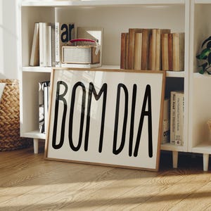 May include: A framed print with the words "BOM DIA" in bold, black letters. The print has a light background and a wooden frame. The print is on a wooden floor in front of a white bookshelf with books and decorative items.