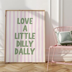 May include: A framed print with a vertical striped pattern in light pink and cream. The text "LOVE A LITTLE DILLY DALLY" is in green. A pink metal chair with a decorative pillow is next to the print.