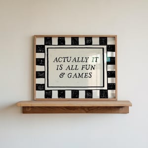 K&ouml;nnte beinhalten: Ein gerahmter Druck mit dem Text "ACTUALLY IT IS ALL FUN & GAMES" in schwarzer Schrift. Der Rahmen hat einen schwarz-wei&szlig; gestreiften Rand. Der Druck liegt auf einem Holzregal an einer wei&szlig;en Wand.