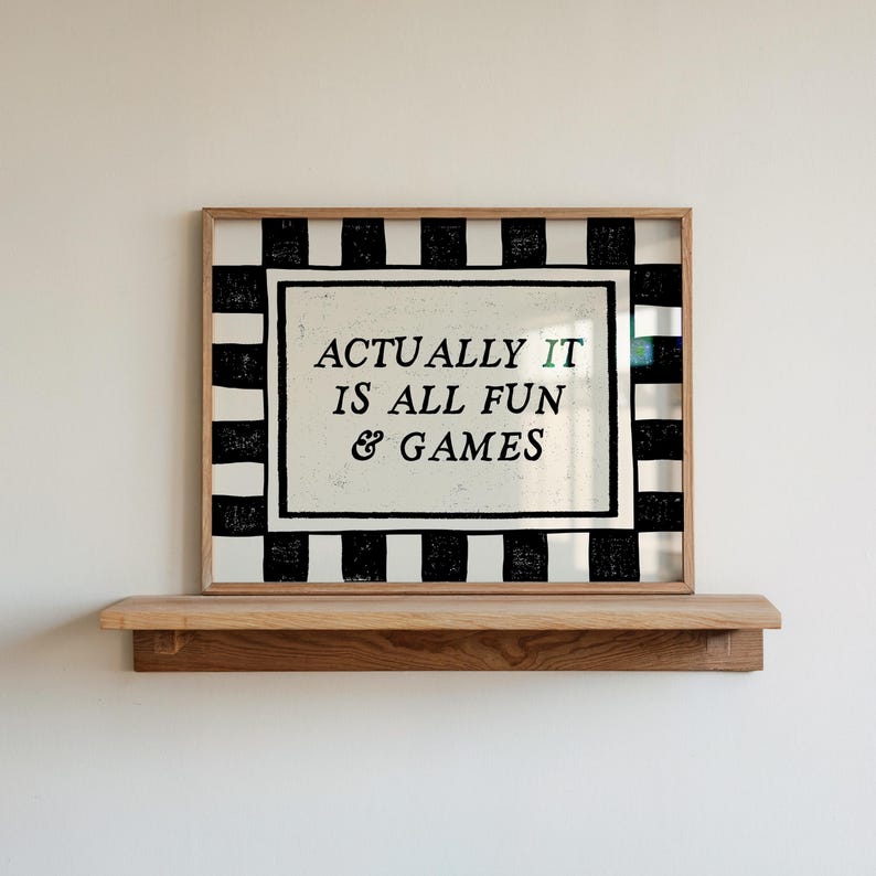 K&ouml;nnte beinhalten: Ein gerahmter Druck mit dem Text "ACTUALLY IT IS ALL FUN & GAMES" in schwarzer Schrift. Der Rahmen hat einen schwarz-wei&szlig; gestreiften Rand. Der Druck liegt auf einem Holzregal an einer wei&szlig;en Wand.