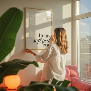 Puede incluir: Un estampado enmarcado con el texto "In my soft girl era" en un marco dorado. El estampado se cuelga en una pared blanca. La imagen est&aacute; en una habitaci&oacute;n bien iluminada con una silla rosa y grandes ventanas.