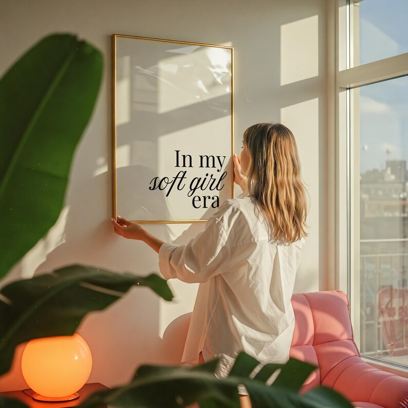 Puede incluir: Un estampado enmarcado con el texto "In my soft girl era" en un marco dorado. El estampado se cuelga en una pared blanca. La imagen est&aacute; en una habitaci&oacute;n bien iluminada con una silla rosa y grandes ventanas.
