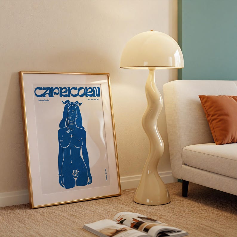 May include: A gold framed poster with a blue illustration of a woman with horns and the word "Capricorn" in blue lettering. The poster is hanging on a white wall next to a white lamp with a white shade.