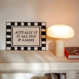 K&ouml;nnte beinhalten: Eine wei&szlig;e Tischlampe mit pilzf&ouml;rmigem Schirm und zylindrischem Sockel. Daneben ein gerahmtes Schild mit dem Text "ACTUALLY IT IS ALL FUN & GAMES" und ein Buch mit rotem Einband.
