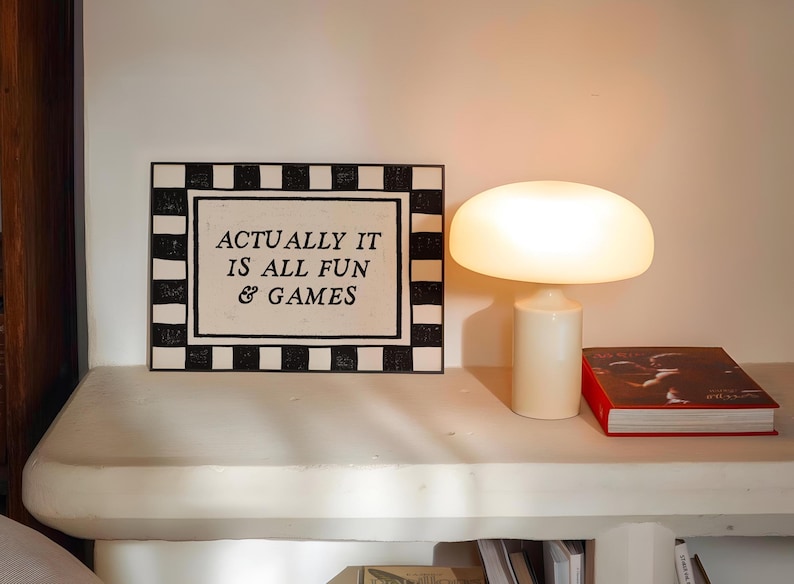 K&ouml;nnte beinhalten: Eine wei&szlig;e Tischlampe mit pilzf&ouml;rmigem Schirm und zylindrischem Sockel. Daneben ein gerahmtes Schild mit dem Text "ACTUALLY IT IS ALL FUN & GAMES" und ein Buch mit rotem Einband.