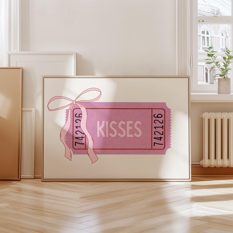May include: A pink ticket-style wall art print with the word 'KISSES' in the center. The ticket has the numbers '742126' printed on both ends. A pink ribbon with a bow is attached to the top left corner. The artwork is displayed in a wooden frame.
