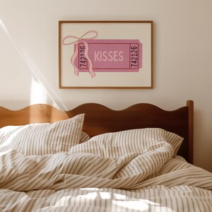 May include: A framed pink ticket art print with the word 'KISSES' in the center. The ticket has a pink ribbon attached to the top left corner. The ticket has the number '742126' printed on both sides. The print is displayed above a bed with striped bedding.
