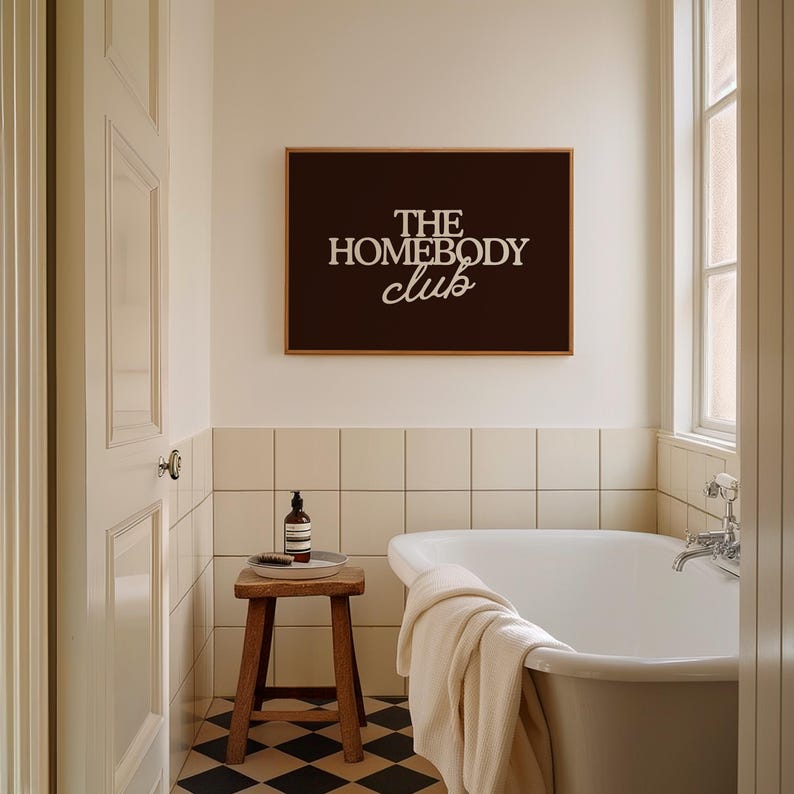 May include: A rectangular wall art print featuring a dark brown background with the white text THE HOMEBODY club centered in a mix of bold serif and script fonts, displayed in a thin wooden frame above a white bathtub and a small wooden stool.