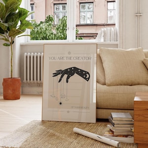 Puede incluir: Un p&oacute;ster beige con una silueta negra de una mano sosteniendo cuerdas con planetas unidos. El texto en el p&oacute;ster dice "T&uacute; eres el creador de tu realidad."