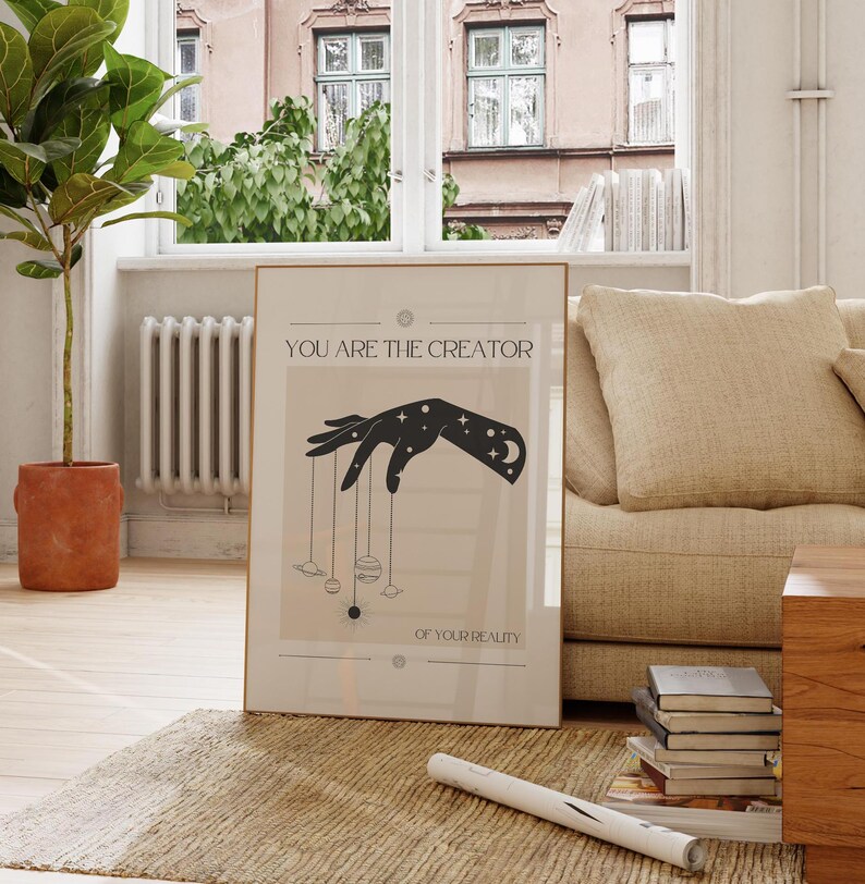 Puede incluir: Un p&oacute;ster beige con una silueta negra de una mano sosteniendo cuerdas con planetas unidos. El texto en el p&oacute;ster dice "T&uacute; eres el creador de tu realidad."