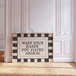 May include: A framed sign with the text "WASH YOUR HANDS YOU FILTHY ANIMAL" in black lettering. The sign has a black and white checkered border and is displayed against a white wall.