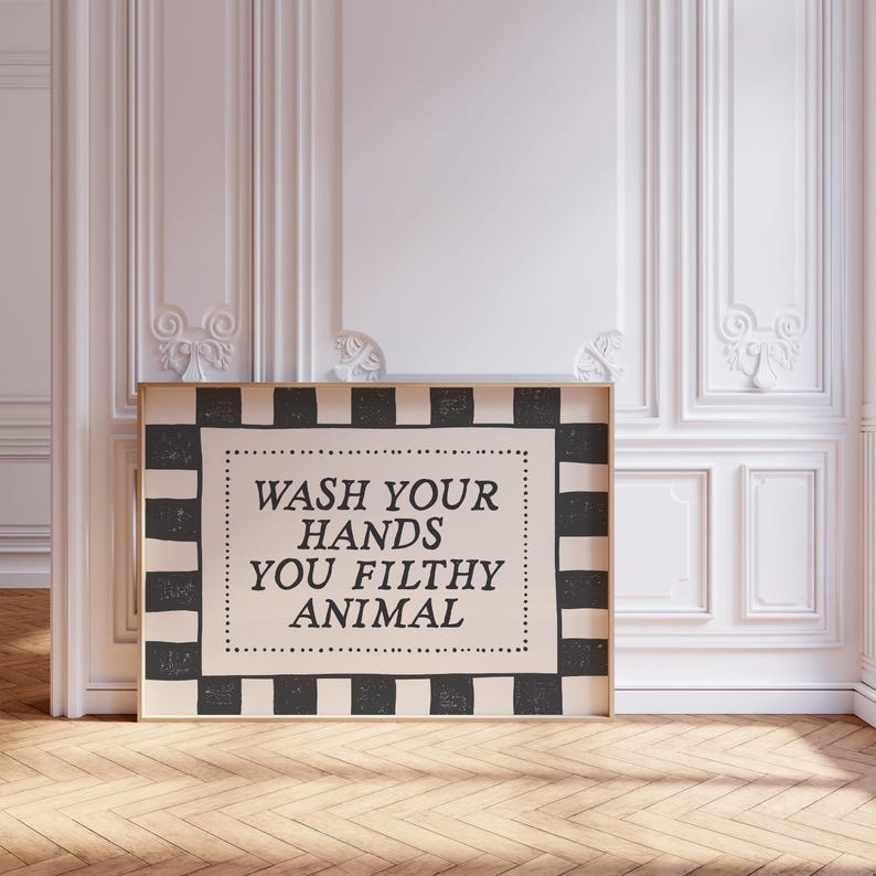 May include: A framed sign with the text "WASH YOUR HANDS YOU FILTHY ANIMAL" in black lettering. The sign has a black and white checkered border and is displayed against a white wall.