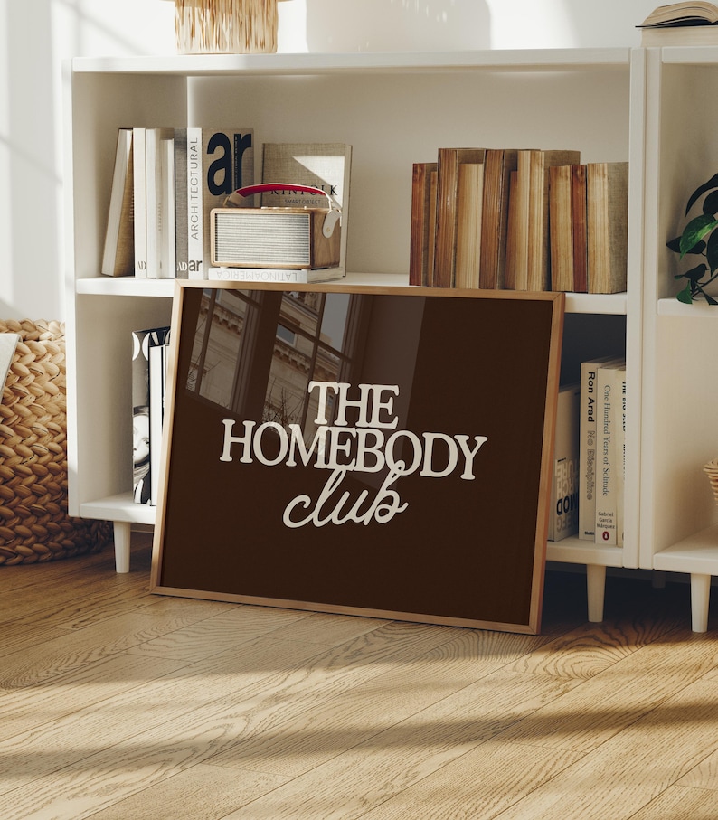 May include: A rectangular framed wall art piece featuring a dark brown background with the white text "THE HOMEBODY club" printed in a mix of bold serif and script fonts. The frame is made of light-colored wood and has a glossy, reflective surface.
