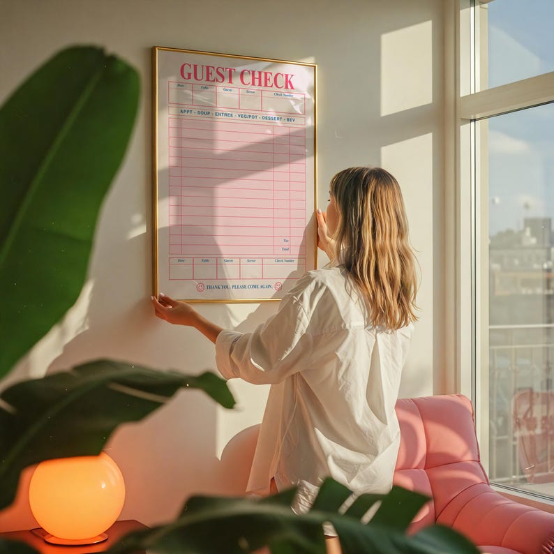 Puede incluir: Un letrero "Guest Check" rosa y dorado se muestra en una pared. El letrero tiene un fondo rosa con l&iacute;neas y texto blancos. El letrero lo sostiene una persona que lleva una camisa blanca. Una silla rosa y una l&aacute;mpara naranja brillante est&aacute;n en primer plano.