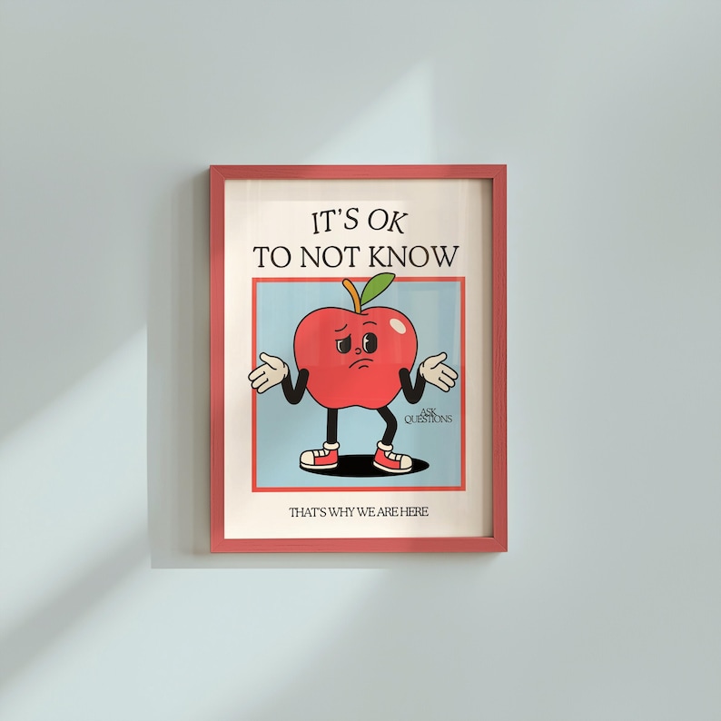 May include: A framed print featuring a cartoon apple character with a sad expression, arms outstretched. The text "IT'S OK TO NOT KNOW" is above the apple, with "THAT'S WHY WE ARE HERE" below. The frame is a reddish-pink color.