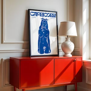 May include: A blue silhouette of a woman with horns and a star on her chest, against a white background. The image is framed in black and the text "CAPRICORN" is at the top of the image.