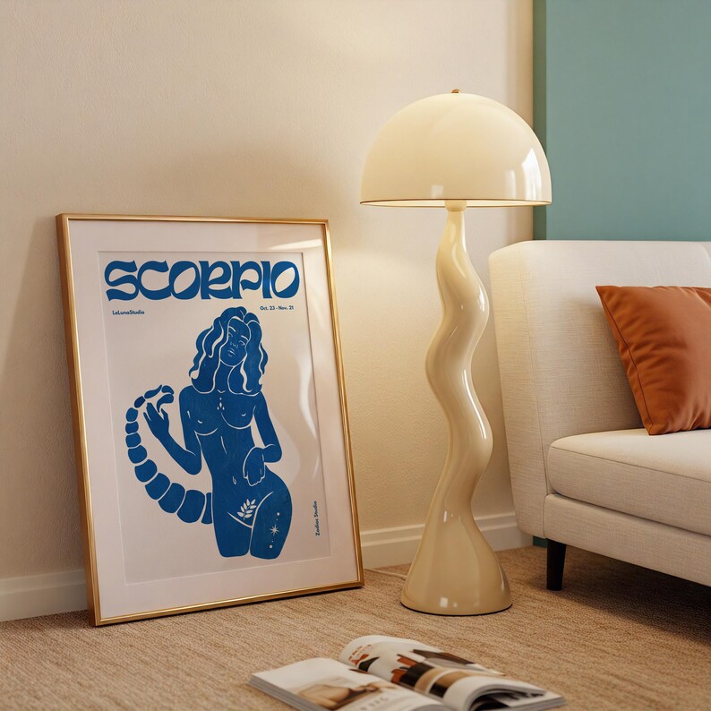 Pode incluir: Um p&ocirc;ster emoldurado em dourado com uma ilustra&ccedil;&atilde;o azul de uma mulher com cauda de escorpi&atilde;o. O p&ocirc;ster tem o texto "Scorpio" em uma fonte azul em negrito. O p&ocirc;ster est&aacute; pendurado em uma parede ao lado de uma l&acirc;mpada branca com um abajur branco.