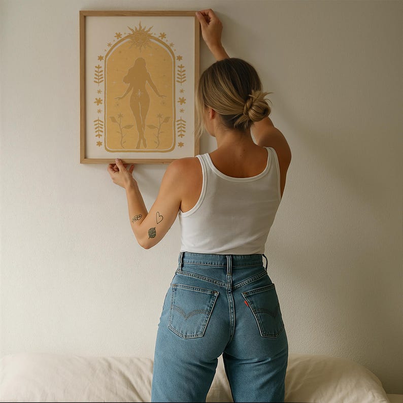 May include: A rectangular framed wall art print featuring a golden yellow illustration of a standing figure centered within an arched border. The design includes a sun motif at the top, star accents, and floral elements on a light cream background.