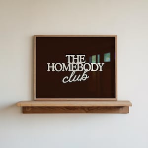 May include: A rectangular framed print with a dark brown background and the white text THE HOMEBODY CLUB centered on the surface. The frame is made of light-colored wood and rests on a matching wooden shelf mounted against a plain wall.