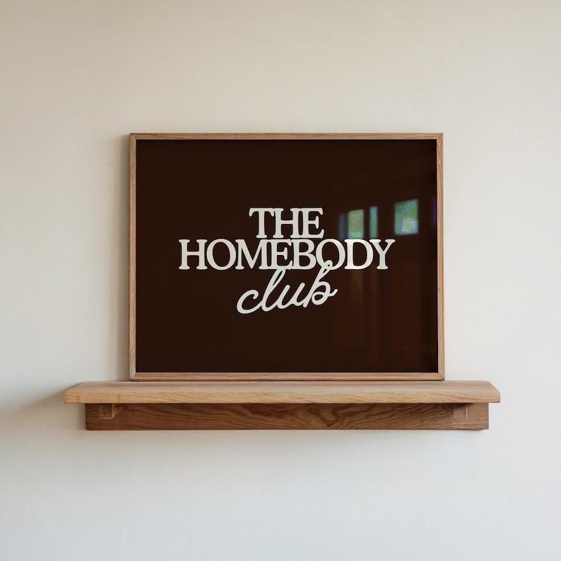 May include: A rectangular framed print with a dark brown background and the white text THE HOMEBODY CLUB centered on the surface. The frame is made of light-colored wood and rests on a matching wooden shelf mounted against a plain wall.