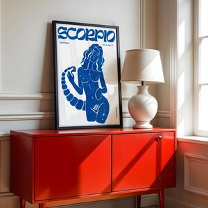 Pode incluir: Uma impress&atilde;o azul e branca de uma mulher com uma cauda de escorpi&atilde;o, com a palavra "Scorpio" em letras negritas no topo. A impress&atilde;o est&aacute; emoldurada em uma moldura preta e est&aacute; pendurada em uma parede branca acima de um arm&aacute;rio vermelho.