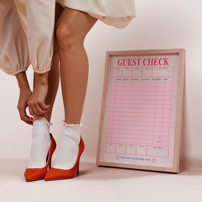 Puede incluir: Un cheque de invitado rosa y blanco enmarcado con las palabras "GUEST CHECK" en la parte superior. El marco es de color marr&oacute;n claro. La imagen tambi&eacute;n muestra un par de tacones altos naranjas y calcetines blancos.