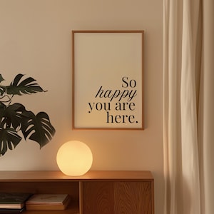 Può includere: Una stampa incorniciata con la scritta "So happy you are here" in nero. Una lampada rotonda e luminosa è appoggiata su un mobile di legno sotto la stampa. Sono visibili anche una grande pianta e una tenda color crema.