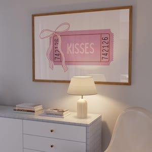 May include: A framed pink 'Kisses' ticket art print. The ticket features the word 'Kisses' in a bold font, with the numbers '742126' on each side. A pink ribbon is tied to the top left corner. The print is displayed above a white cabinet with a marble top, a lamp, and a white chair.