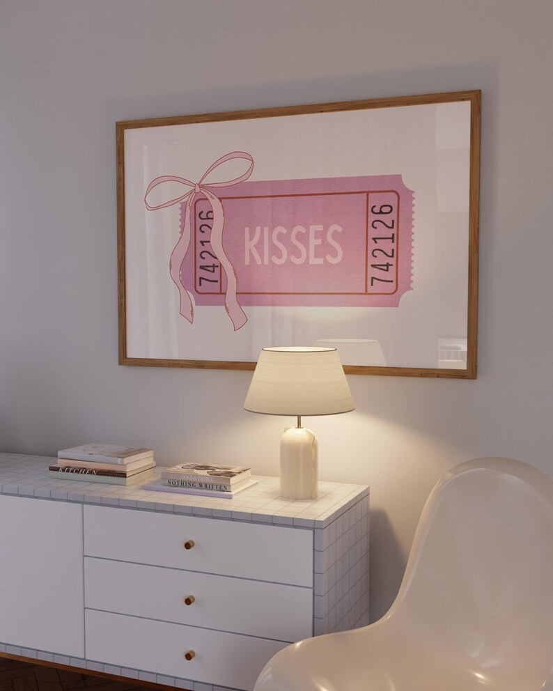 May include: A framed pink 'Kisses' ticket art print. The ticket features the word 'Kisses' in a bold font, with the numbers '742126' on each side. A pink ribbon is tied to the top left corner. The print is displayed above a white cabinet with a marble top, a lamp, and a white chair.