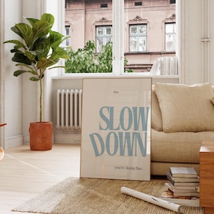 Może przedstawiać: Oprawiony plakat ze słowami "SLOW DOWN" w jasnoniebieskim kolorze, z tekstem "Hey" powyżej i "you're doing fine." poniżej. Plakat opiera się o beżową sofę w pokoju z dużą rośliną i oknem.