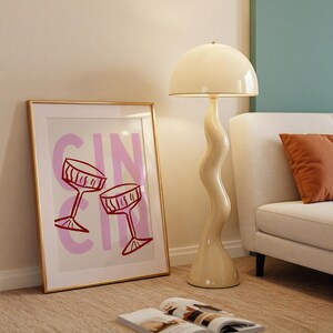 Puede incluir: Un cuadro enmarcado con la palabra "GIN" en rosa y dos copas de c&oacute;ctel rojas. Una l&aacute;mpara de pie crema con un tallo ondulado y una pantalla de c&uacute;pula est&aacute; junto al cuadro. Una revista abierta descansa en el suelo.