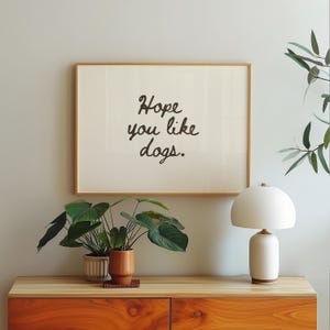May include: A framed print with the text "Hope you like dogs" in black script. The artwork hangs above a wooden cabinet with a white lamp and potted plants. The cabinet has a warm orange finish.