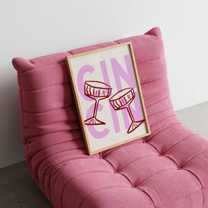 Puede incluir: Una impresi&oacute;n art&iacute;stica enmarcada con la palabra "GIN" en rosa y dos copas de c&oacute;ctel rojas. La impresi&oacute;n descansa sobre un sill&oacute;n de pana rosa. La obra de arte est&aacute; en un marco de madera clara.