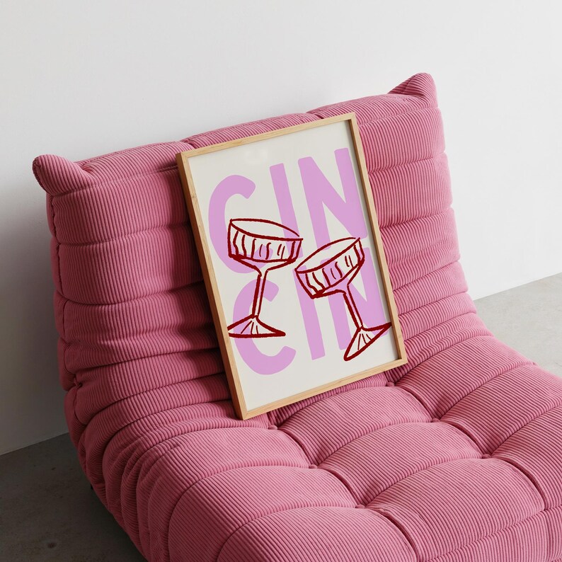 Puede incluir: Una impresi&oacute;n art&iacute;stica enmarcada con la palabra "GIN" en rosa y dos copas de c&oacute;ctel rojas. La impresi&oacute;n descansa sobre un sill&oacute;n de pana rosa. La obra de arte est&aacute; en un marco de madera clara.