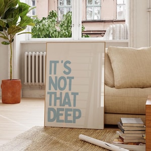 Pode incluir: Um poster emoldurado com o texto "IT'S NOT THAT DEEP" em letras azul claro. O poster é exibido numa sala com uma janela, um sofá bege e um tapete tecido. Uma planta em vaso e uma pilha de livros também são visíveis.