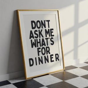 Pode incluir: Uma impressão emoldurada com o texto "DON'T ASK ME WHAT'S FOR DINNER" em negrito preto. A impressão tem um fundo branco e está em uma moldura dourada. Está encostada em uma parede branca, com um chão de azulejos preto e branco.