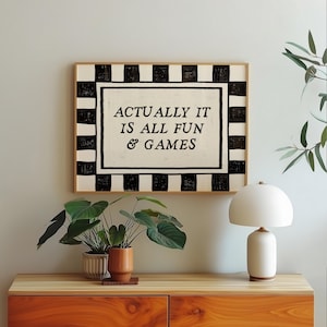 K&ouml;nnte beinhalten: Ein gerahmter Kunstdruck mit dem Text "ACTUALLY IT IS ALL FUN & GAMES" in einem schwarz-wei&szlig;en Schachbrettmuster. Das Kunstwerk wird &uuml;ber einem Holzschrank mit Topfpflanzen und einer wei&szlig;en Tischlampe pr&auml;sentiert.