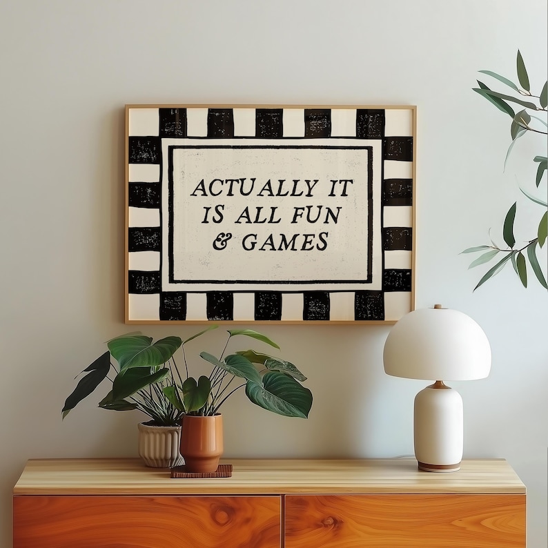 K&ouml;nnte beinhalten: Ein gerahmter Kunstdruck mit dem Text "ACTUALLY IT IS ALL FUN & GAMES" in einem schwarz-wei&szlig;en Schachbrettmuster. Das Kunstwerk wird &uuml;ber einem Holzschrank mit Topfpflanzen und einer wei&szlig;en Tischlampe pr&auml;sentiert.