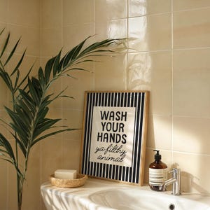 May include: A bathroom scene featuring a framed sign with the text "WASH YOUR HANDS ya filthy animal". The sign has a black and white striped border. A small woven basket holds a bar of soap. A brown soap bottle and a silver tap are also visible.