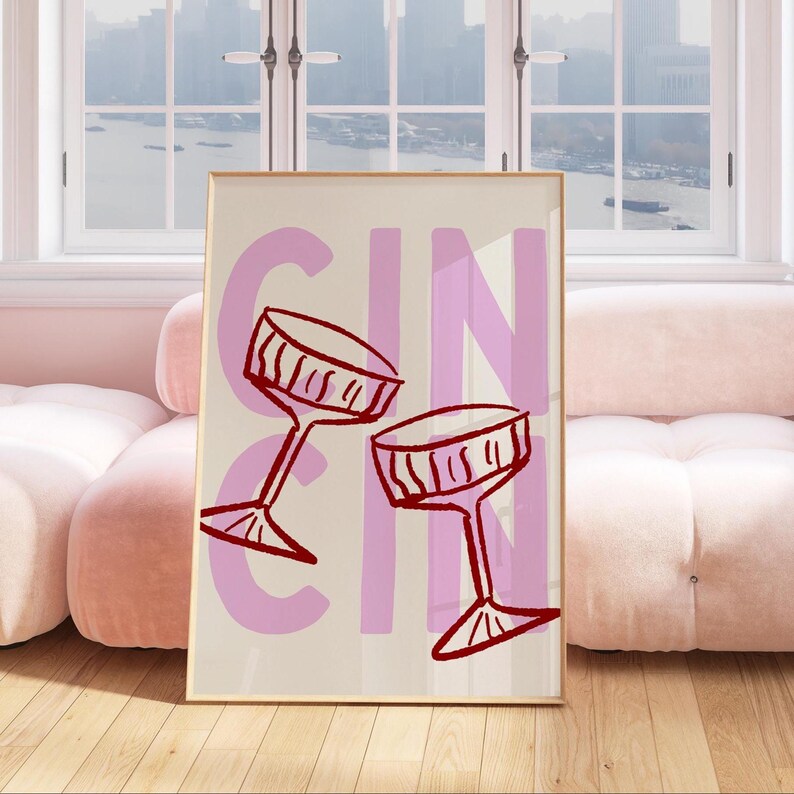 Puede incluir: Un cuadro enmarcado con la palabra "GIN" en rosa y dos copas de c&oacute;ctel rojas estilizadas. El fondo es de color beige claro. El cuadro se exhibe en una habitaci&oacute;n con un sof&aacute; rosa y suelo de madera.