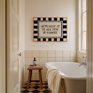 K&ouml;nnte beinhalten: Badezimmer-Interieur mit wei&szlig;er Badewanne, Holzschemel und gerahmtem Kunstwerk. Das Kunstwerk zeigt den Text "ACTUALLY IT IS ALL FUN & GAMES" in einem schwarz-wei&szlig; karierten Rahmen. Der Boden hat ein schwarz-wei&szlig; kariertes Muster.