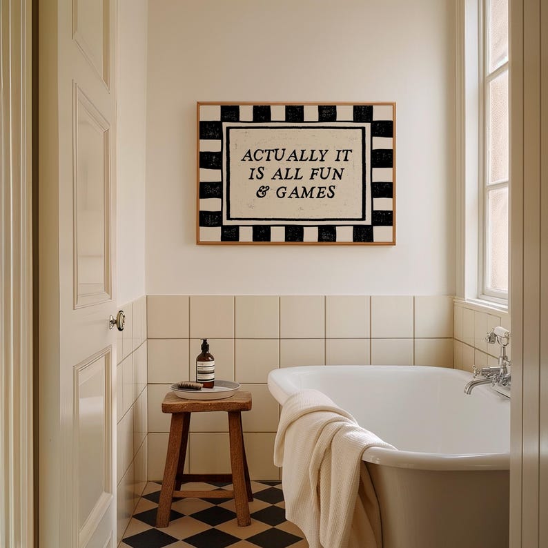 K&ouml;nnte beinhalten: Badezimmer-Interieur mit wei&szlig;er Badewanne, Holzschemel und gerahmtem Kunstwerk. Das Kunstwerk zeigt den Text "ACTUALLY IT IS ALL FUN & GAMES" in einem schwarz-wei&szlig; karierten Rahmen. Der Boden hat ein schwarz-wei&szlig; kariertes Muster.