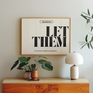 May include: A framed print with the words "LET THEM" in large black letters. Above the text is the word "Reminder" and below it reads "Everyones on their own journey." The print hangs above a wooden cabinet with plants and a white lamp.