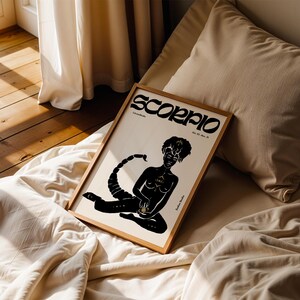 May include: A black and white poster with the word "Scorpio" in a stylized font. The poster features a black silhouette of a person with a scorpion tail, holding a small circle in their hand. The poster is framed in a light wood frame and is lying on a bed with white linens.