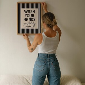 May include: A framed print with the text "WASH YOUR HANDS ya filthy animal". The artwork has a black and white striped border. The person holding the print is wearing a white vest top and blue jeans.