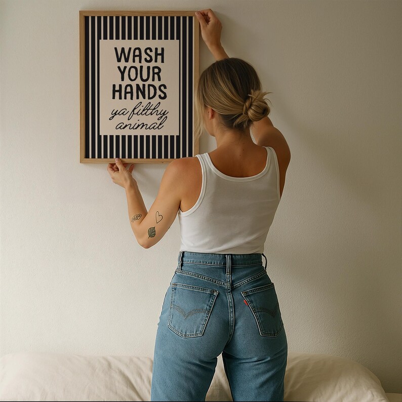 May include: A framed print with the text "WASH YOUR HANDS ya filthy animal". The artwork has a black and white striped border. The person holding the print is wearing a white vest top and blue jeans.