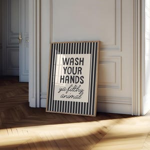 May include: A framed bathroom print with a black and white striped border. The print features the text "WASH YOUR HANDS ya filthy animal" in a bold, black font on a white background. The frame is a light brown colour.