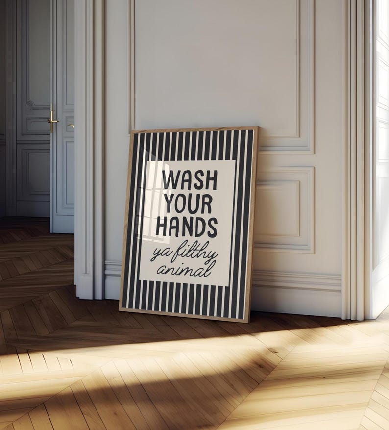 May include: A framed bathroom print with a black and white striped border. The print features the text "WASH YOUR HANDS ya filthy animal" in a bold, black font on a white background. The frame is a light brown colour.
