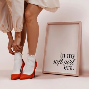 Puede incluir: Un par de tacones altos naranjas con calcetines blancos acanalados y una impresi&oacute;n enmarcada. La impresi&oacute;n dice "In my soft girl era." Los tacones tienen una punta puntiaguda y un tac&oacute;n de aguja. Los calcetines tienen un borde con volantes.