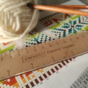 Puede incluir: Un primer plano de una regla de madera con las palabras "JENERATES Pattern Tracker" impresas en ella. La regla se coloca sobre un patrón de cuadrícula blanco y de colores. También se puede ver una bola de hilo blanco y dos agujas de tejer de madera en la imagen.